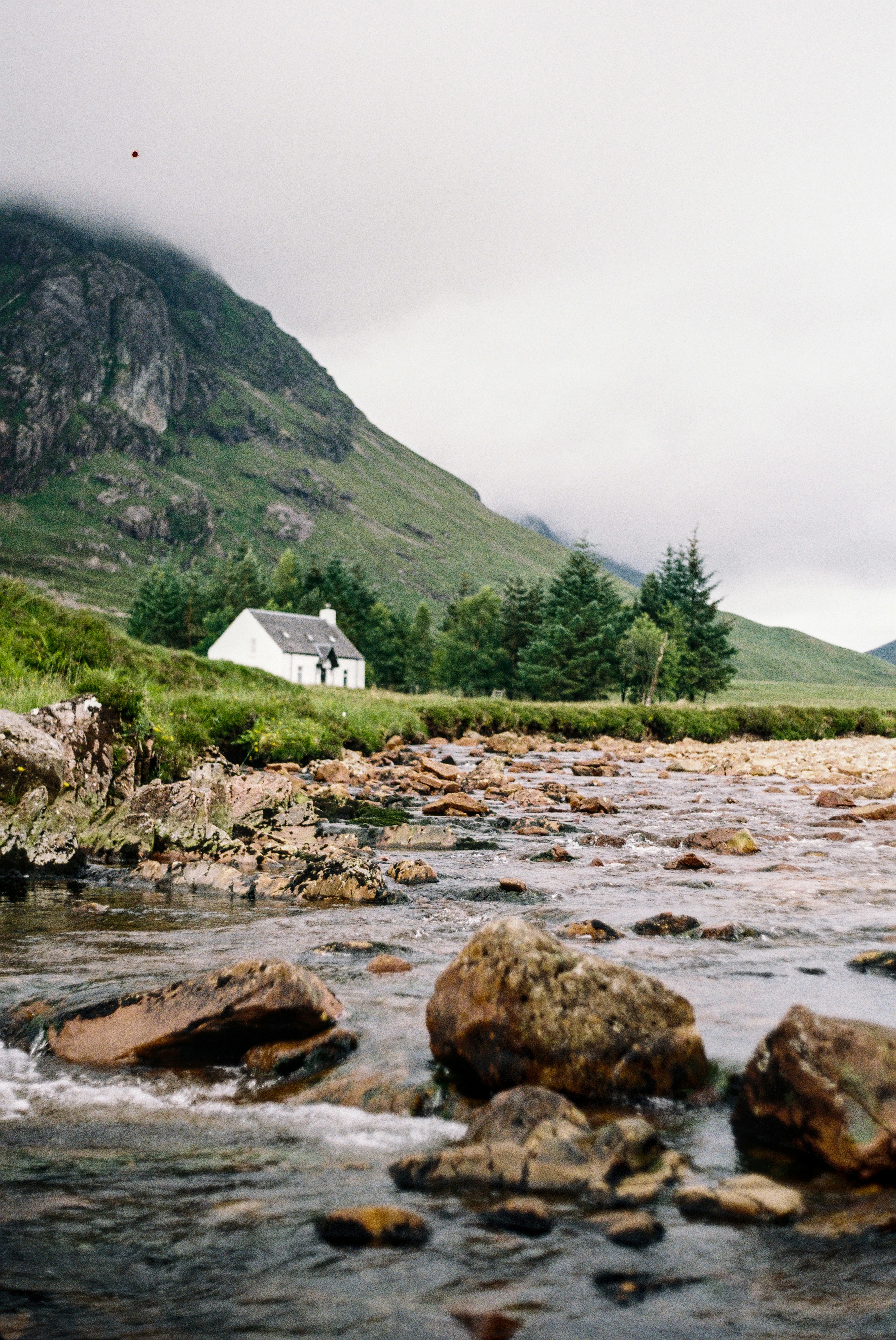 En vit liten stuga som ligger jämte en bäck omringat av skogsklädda berg 